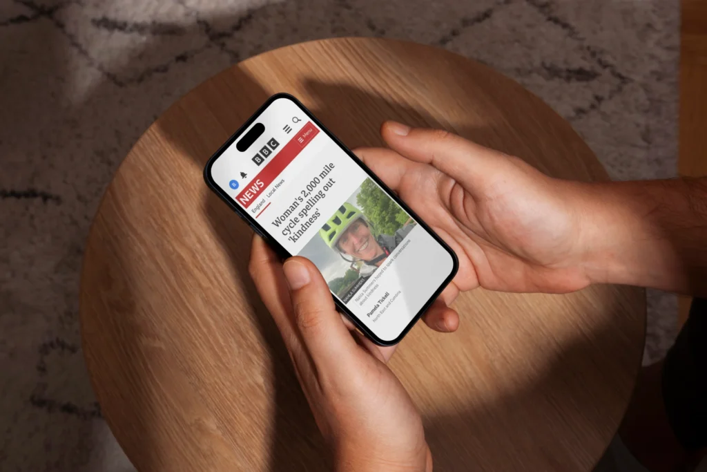 Hands hold a smartphone displaying a news article above a round wooden table.