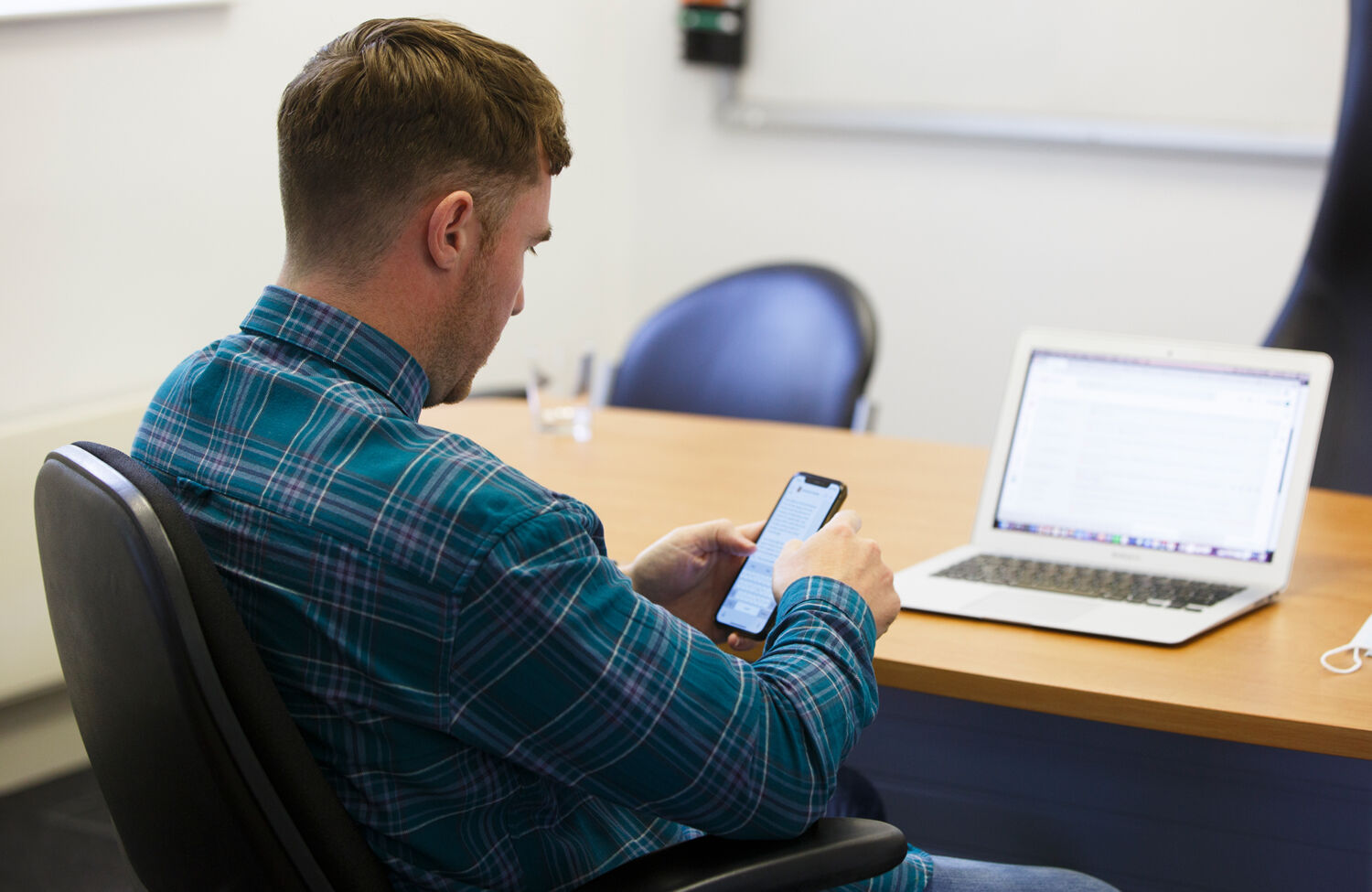 Man seated at a desk using a smartphone beside an open laptop in an office
