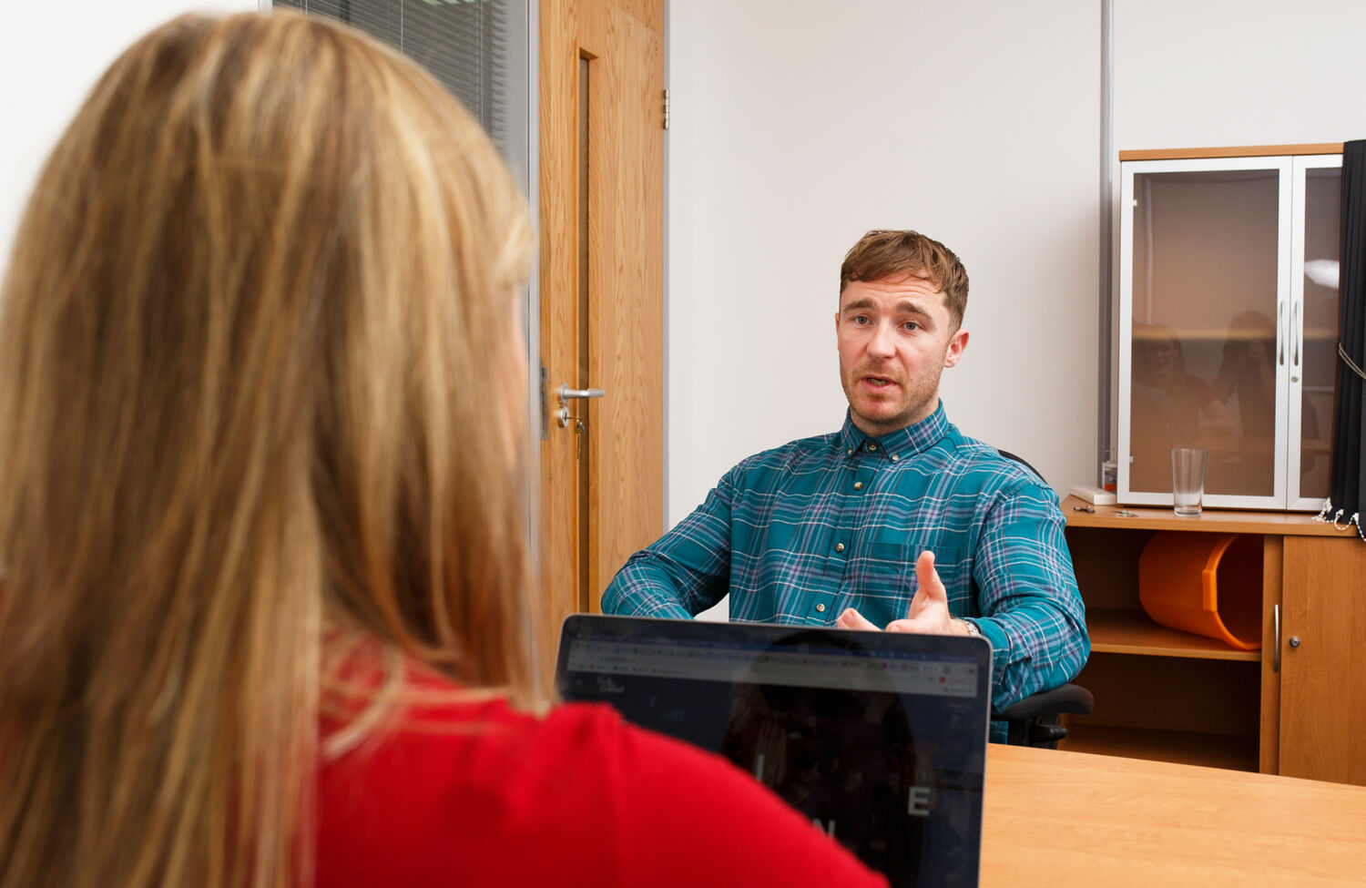 Man talks with a woman across a desk in an office.
